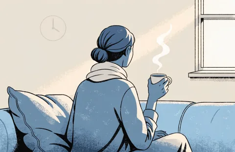 A person seen from behind, sitting on a sofa with a warm cup and a light scarf around the neck, by a window.